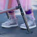 A close-up of a child's feet wearing sparkly silver high-top sneakers with white soles, pink patterned socks, and a pink tutu skirt, standing near a metal walker outdoors.